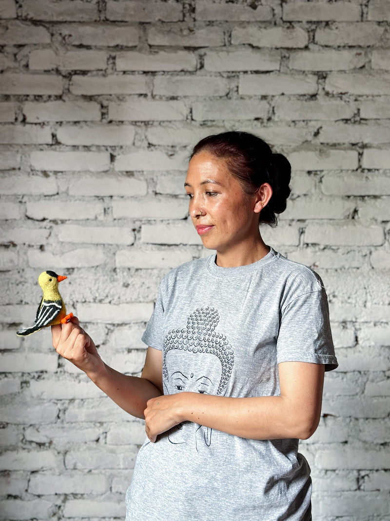 A handmade needle-felted Goldfinch finger puppet is presented on the fingertip of a Nepalese female artisan, set against a white brick wall. This tiny wool felt goldfinch puppet features a vivid yellow body, black head, orange beak, and black and white wings with delicate hand-felted details. Ethically crafted, it supports female artisans in Nepal.