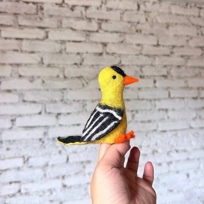 A handcrafted wool felt goldfinch finger puppet is held by a hand against a blurred white brick wall. This vibrant needle-felted goldfinch puppet features a bright yellow body, black head marking, orange beak and feet. Its black wings have white stripes, and the tail is tipped with black, highlighting the textures of this ethically made felt toy.