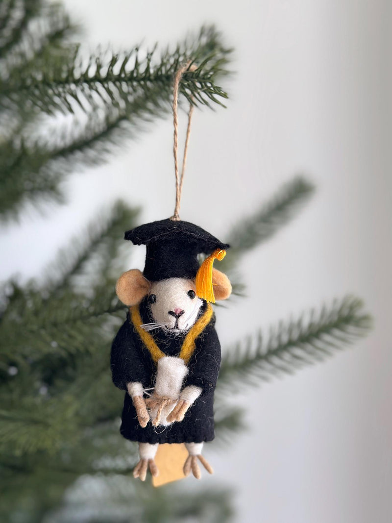 A needle felted wool felt Graduation Mouse Ornament hangs in a close-up against a soft green pine and white background. The white mouse, with pink nose and ear details, is adorned in a black graduation robe and cap with a yellow tassel, clutching a tiny tan diploma. A brown jute string for hanging is attached to the cap.