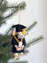 A wool felt graduation mouse ornament hangs from a jute string against a blurred background of evergreen branches and a white wall. This handmade ornament features a white mouse in a black graduation robe and cap with a yellow tassel, holding a tiny diploma. It has wire legs, hands, a tail, small round ears, and a pink nose, perfect for commemorating academic achievements.