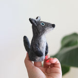 A handmade, needle-felted gray coyote finger puppet is held up by a female hand with red nail polish, against a bright, blurred green background. This unique wool felt finger puppet features a gray body, lighter gray belly, dark gray paws, perky ears, a pointed snout with a black nose, and round blue eyes.