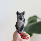 A handmade needle felted gray coyote finger puppet is worn on a female hand with red nail polish, set against a soft white background with a hint of greenery. This handcrafted wool felt toy is predominantly gray with darker gray paws and inner ears. It features bright blue eyes, a small black nose, and a visible small tail on its back, showcasing its textured needle-felted appearance.