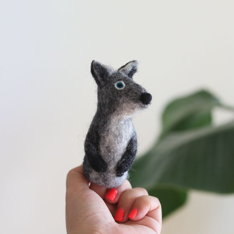 A handmade needle felted gray coyote finger puppet is playfully held by a female hand with red-painted nails, set against a bright backdrop with blurred green foliage. This handcrafted wool felt finger puppet showcases gray and white felt, small perky ears, a rounded black nose, and bright blue eyes, highlighting its unique design.
