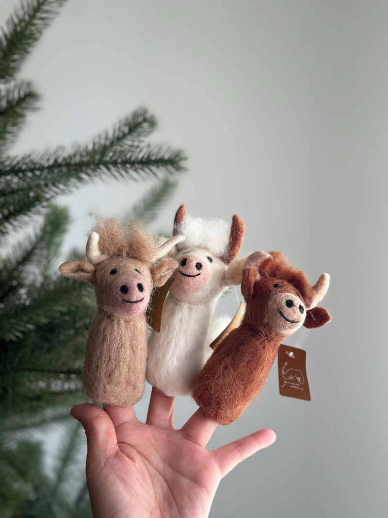 A cheerful wool felt Highland Cow finger puppet is displayed on a female hand, with a festive Christmas tree subtly blurred in the background. This charming, handmade felt finger puppet comes in light brown, white, and brown hues, each featuring distinct horns and a smiling face, perfect for engaging storytelling.