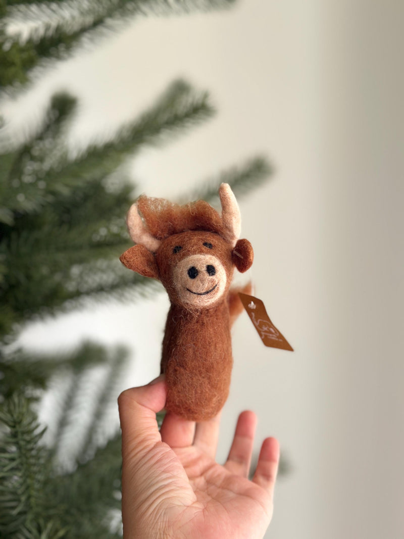 A playful, needle felted Highland cow finger puppet is held by a female hand, presented against a blurred Christmas tree background. This charming, handmade felt toy features brown fluffy hair, small ears, and curved horns, with black dot eyes and a stitched smile. A small tag is attached to its tail.