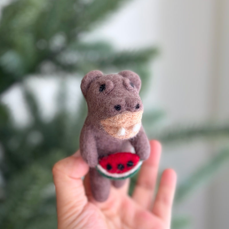 A handcrafted felt hippopotamus finger puppet, holding a watermelon slice, is delicately presented by a female hand in a bright, airy setting. This muted brown hippopotamus features small, rounded ears and tiny black eyes. The vibrant red watermelon slice it holds is detailed with black seeds and a thin green rind. Blurred greenery in the background suggests a playful, summery atmosphere.