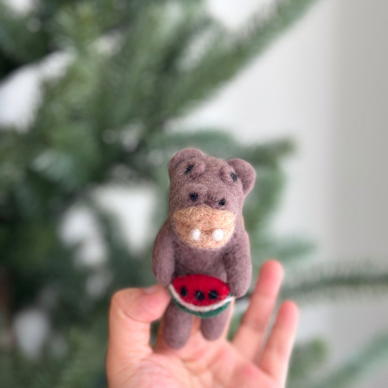 A light brown wool felt hippopotamus holding watermelon finger puppet is held in a female hand, set against a blurred Christmas pine tree. This cheerful handcrafted hippo features round ears and two small white tusks. It playfully clutches a red watermelon slice with black seeds and a green rind, embodying a joyful summer theme for imaginative play.