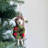 A wool felt sheep ornament hangs on a green Christmas tree branch against a soft white background. The sheep has a soft gray, textured body, a white face with small white ears, and a stitched black 'X' for its mouth. It holds a textured green holly wreath adorned with three red berries. A jute string is attached for hanging this charming Christmas ornament.