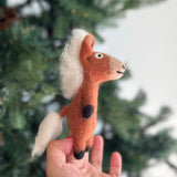 A handmade wool felt horse finger puppet is presented by a female hand, set against a softly blurred Christmas tree background. This charming light brown horse features a cream mane and tail, black hooves, and expressive black eyes with stitched details. Crafted from 100% wool felt and meticulously needle-felted, this unique kids' finger puppet is ideal for imaginative play and makes a lovely holiday gift.