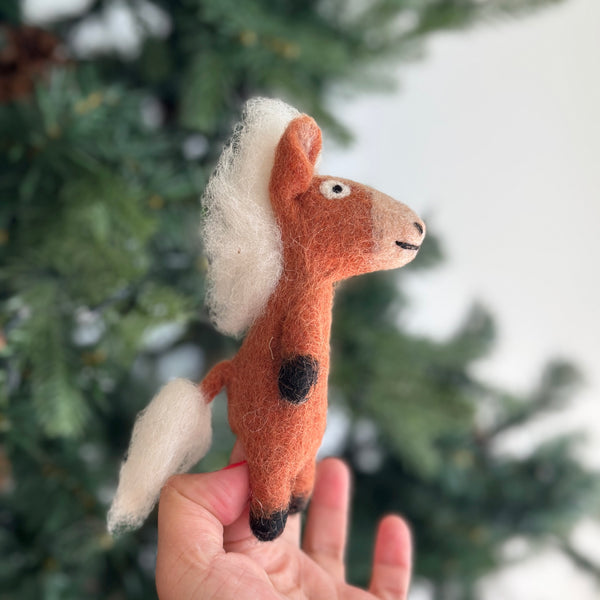 A handmade wool felt horse finger puppet is presented by a female hand, set against a softly blurred Christmas tree background. This charming light brown horse features a cream mane and tail, black hooves, and expressive black eyes with stitched details. Crafted from 100% wool felt and meticulously needle-felted, this unique kids' finger puppet is ideal for imaginative play and makes a lovely holiday gift.