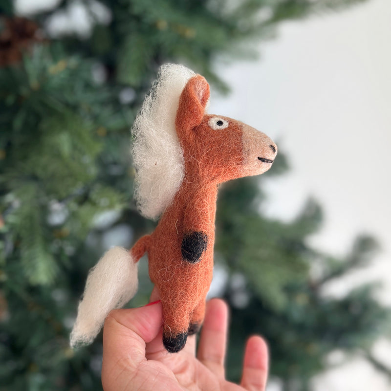 A handmade wool felt horse finger puppet is presented by a female hand, set against a softly blurred Christmas tree background. This charming light brown horse features a cream mane and tail, black hooves, and expressive black eyes with stitched details. Crafted from 100% wool felt and meticulously needle-felted, this unique kids' finger puppet is ideal for imaginative play and makes a lovely holiday gift.