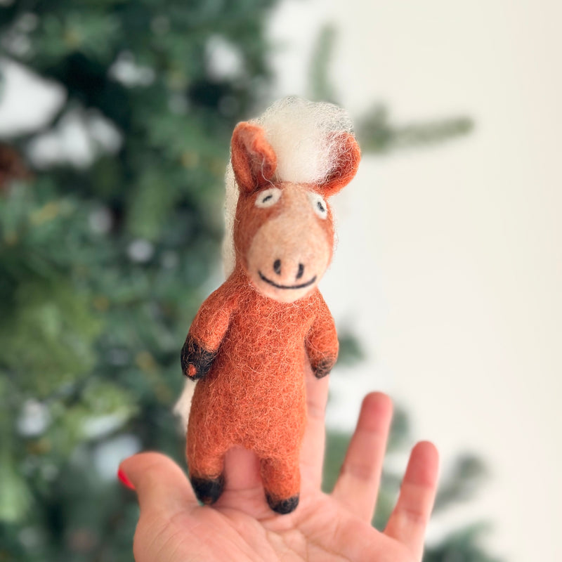 A handmade wool felt horse finger puppet is presented on a female hand, set against a blurred Christmas tree background, creating a festive scene. This reddish-brown needle felted horse puppet features a fluffy white mane, black hooves, and a charming stitched smile. Crafted from 100% wool in Nepal, it highlights artisanal skill and fair-trade principles, perfect for storytelling.