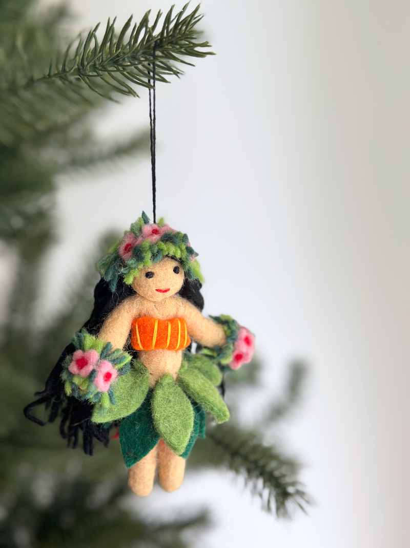 A festive felt hula girl ornament hangs on a Christmas tree against a light background. This unique handmade ornament features a smiling, light brown hula girl with black yarn hair, wearing a green leaf skirt, an orange top, and a green flower headpiece adorned with pink flowers. Matching flower bracelets complete her look, and a black string allows for hanging.