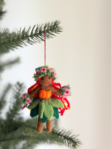 A handcrafted Hula Girl felt ornament hangs gracefully from a pine branch against a soft white background. This unique Christmas ornament features brown skin, a floral headpiece, and a leafy green grass skirt. Bright orange accents adorn her top, with colorful red and yellow strands suggesting hair, adding tropical charm.