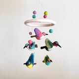 A felt Hummingbird Baby Mobile is suspended against a plain, light background, showcasing its vibrant design. Five colorful felt hummingbirds, each with unique wing poses, hang from a white felt frame by thin white strings. The birds feature appealing color combinations, including purple and pink, green and white, and various shades of blue and yellow. This handcrafted mobile offers a charming and visually engaging experience.
