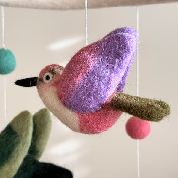 A felt hummingbird from the Hummingbird Baby Mobile is shown in a close-up against a soft, light background. Suspended by thin white strings, this vibrant bird features dynamic wings in purple and pink textured felt. Its body is white and pink, with a black beak and moss-green tail. Parts of the white felt mobile frame, teal and pink felt balls, and other green and black felt hummingbirds are also visible.
