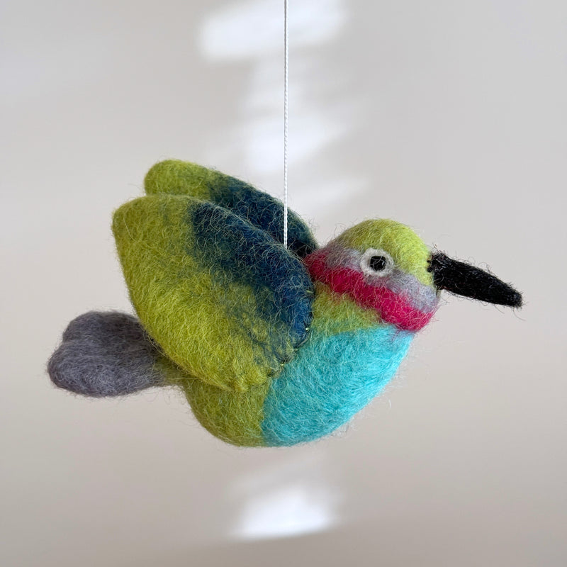 A vibrant wool felt hummingbird, part of a Hummingbird Baby Mobile, is suspended from a white felt frame by a thin white string against a light gray backdrop. This colorful felt hummingbird features a black beak, light green head, a pink and gray eye stripe, a blue belly, and green wings with a blue gradient, ending in a small gray tail. The soft felt material gives it a textured appearance.