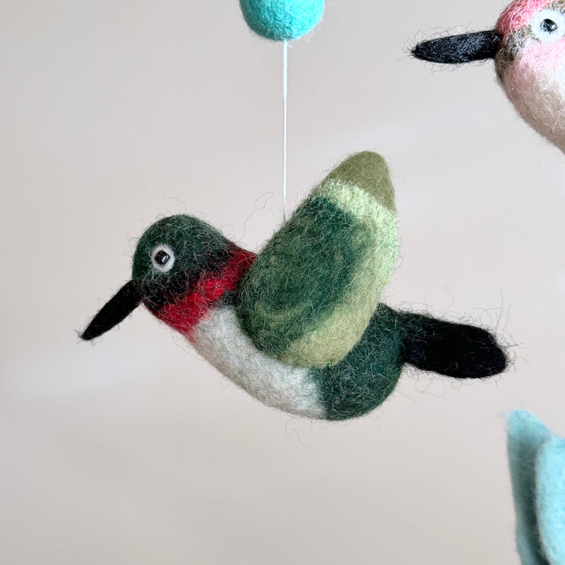 A close-up view of a whimsical Hummingbird felt baby mobile highlights its delicate handcrafted details against a soft, neutral background. A white felt frame holds a suspended wool felt hummingbird, featuring a green head, red throat, white body, and black beak. Above it, a turquoise spherical felt ball hangs by a white string. A second hummingbird is partially visible on the right.