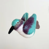 A vibrant felt hummingbird from the Hummingbird Baby Mobile is suspended mid-air against a plain background. This felt hummingbird features a long black beak and a small eye. Its head and wings are turquoise felt, blending into purple tips, with a white felt underside, creating an illusion of flight.