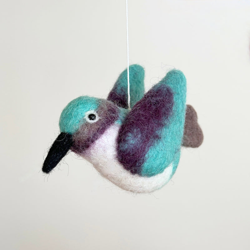 A vibrant felt hummingbird from the Hummingbird Baby Mobile is suspended mid-air against a plain background. This felt hummingbird features a long black beak and a small eye. Its head and wings are turquoise felt, blending into purple tips, with a white felt underside, creating an illusion of flight.