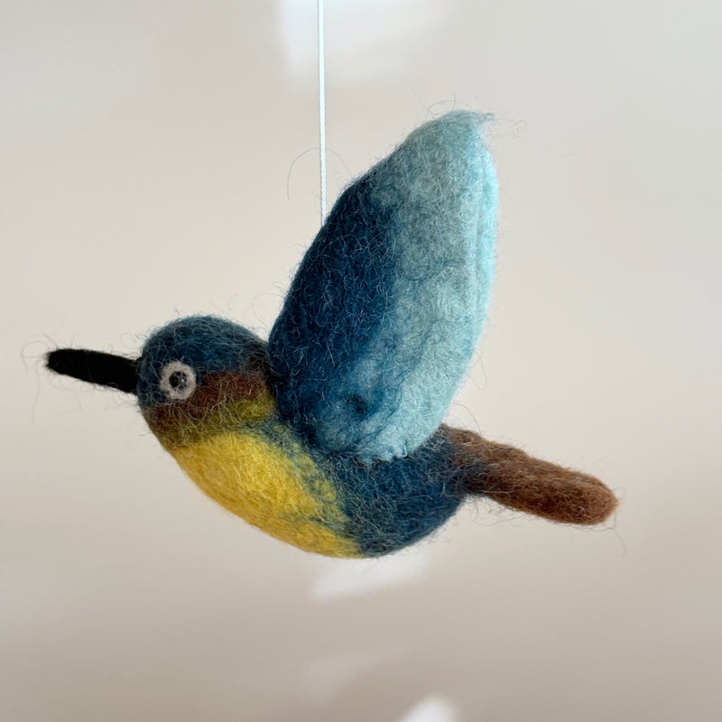 A vibrant wool felt hummingbird, a key element of the Hummingbird Baby Mobile, is shown in close-up, suspended by a thin white string against a muted backdrop, creating the impression of flight. This handcrafted hummingbird features a blue head, yellow chest, and brown tail, with dynamically raised, blue-shaded wings. A long black beak and a small white-and-brown eye add to its lifelike appearance. Its soft, slightly fuzzy felt texture conveys warmth and handmade quality, ideal for a comforting nursery.