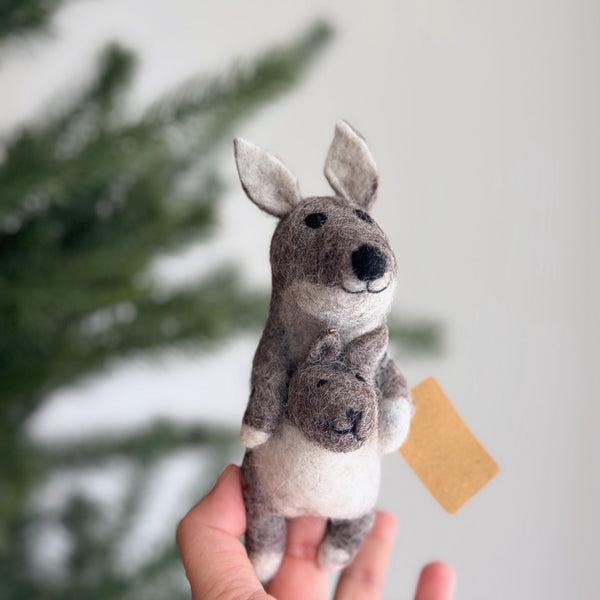 A handcrafted wool felt kangaroo finger puppet is gently held by a hand, set against a soft, neutral backdrop with a blurred green pine branch. This detailed needle-felted puppet features a light brown and gray mama kangaroo with a cream belly and black eyes, carrying a smaller baby kangaroo in its pouch. Handmade from 100% wool felt, this fair-trade felt toy exemplifies artisanal craftsmanship.