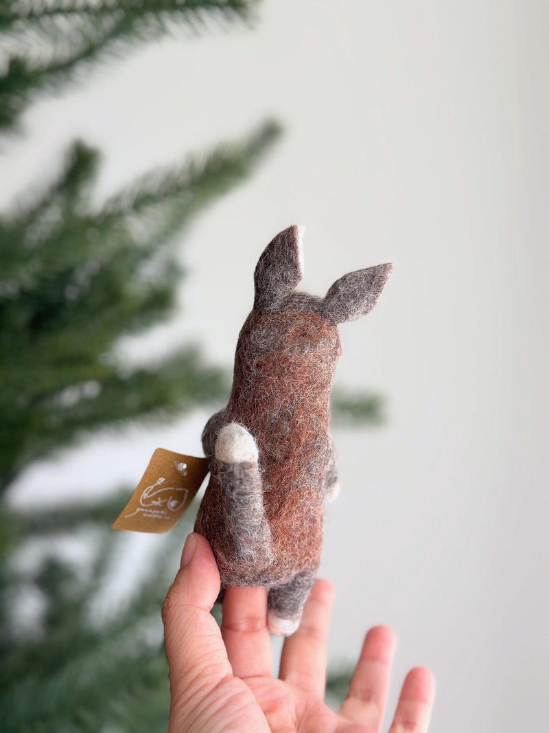A brown wool felt kangaroo finger puppet is held in a hand, set against a soft, bright background with a blurred Christmas pine tree. This handcrafted puppet features pointed ears, a small tail, and lighter accents on its paws and ear insides, highlighting its charming, tactile design and eco-friendly craftsmanship. A small tag hangs from it, emphasizing its handmade origin.