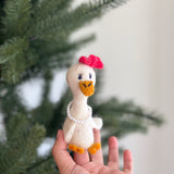 A handmade 100% wool felt Lady Duck finger puppet is presented by a female hand against a blurred Christmas pine tree backdrop. This needle-felted toy is white with a textured orange beak and feet. It is adorned with a pink bow on its head, a small pearl necklace, and features embroidered blue eyes with lashes, highlighting its unique design.