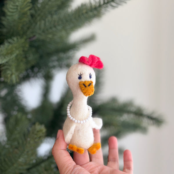A handmade 100% wool felt Lady Duck finger puppet is presented by a female hand against a blurred Christmas pine tree backdrop. This needle-felted toy is white with a textured orange beak and feet. It is adorned with a pink bow on its head, a small pearl necklace, and features embroidered blue eyes with lashes, highlighting its unique design.