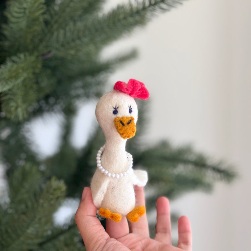 A handmade 100% wool felt Lady Duck finger puppet is presented by a female hand against a blurred Christmas pine tree backdrop. This needle-felted toy is white with a textured orange beak and feet. It is adorned with a pink bow on its head, a small pearl necklace, and features embroidered blue eyes with lashes, highlighting its unique design.