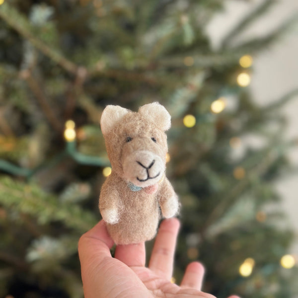 A handcrafted wool felt llama finger puppet is gently held by a female hand, set against a softly blurred Christmas tree background with twinkling lights. This light brown llama puppet features white ears, a cute smile, a soft, fuzzy texture, and a pastel pompom collar, creating a cozy and festive holiday scene.