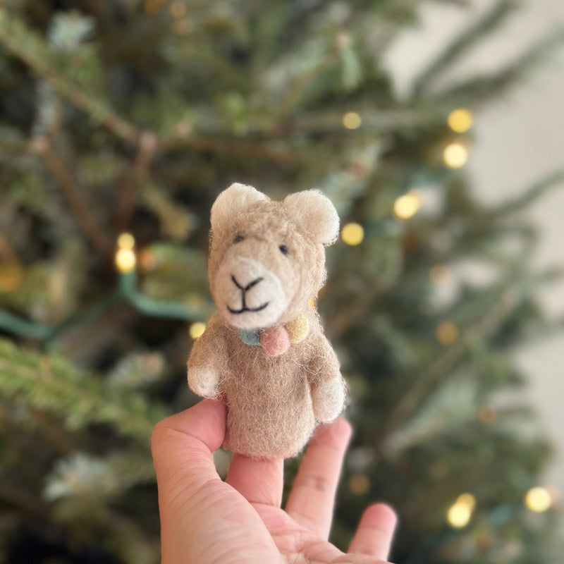 A handcrafted felt llama finger puppet is gently held by a female hand, appearing against a softly blurred Christmas tree with warm lights. The light brown llama features a sweet smile, soft fuzzy texture, white ear and foot details, and a charming pastel pompom collar.