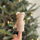 A handmade needle felted llama finger puppet is displayed on a female hand, set against the soft glow of a Christmas tree adorned with warm lights. This handcrafted llama features a neutral tan color, fuzzy texture, rounded ears, a cute smile, and a pastel pompom ball collar decoration.