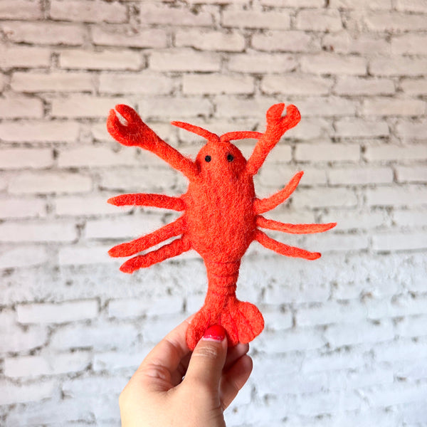 A vibrant red wool felt lobster finger puppet, a unique handcrafted kids toy made through fair-trade practices, is held upright by a female hand against a light grey brick wall. This handmade felt puppet features black eyes, two antennae, six legs, and two prominent claws, showcasing its detailed craftsmanship.