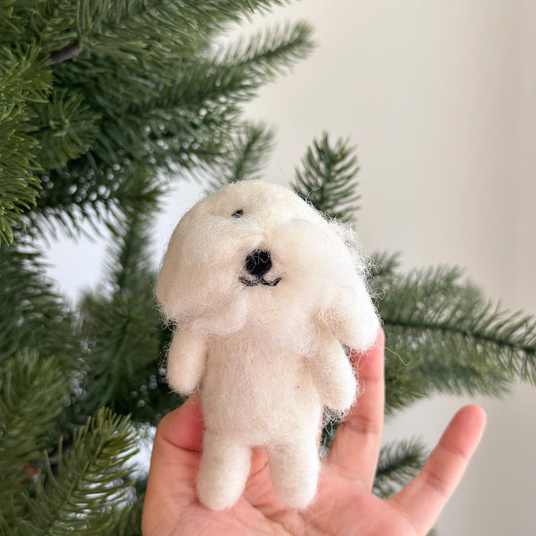 A handcrafted needle felted Maltese dog finger puppet is positioned in front of a subtly blurred Christmas tree. This white wool felted finger puppet features fluffy white wool, a small black nose, and tiny black eyes, capturing the cute and cuddly essence of the pet with lifelike details.