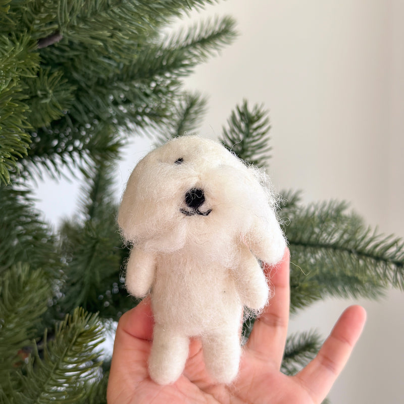 A handcrafted needle felted Maltese dog finger puppet is positioned in front of a subtly blurred Christmas tree. This white wool felted finger puppet features fluffy white wool, a small black nose, and tiny black eyes, capturing the cute and cuddly essence of the pet with lifelike details.