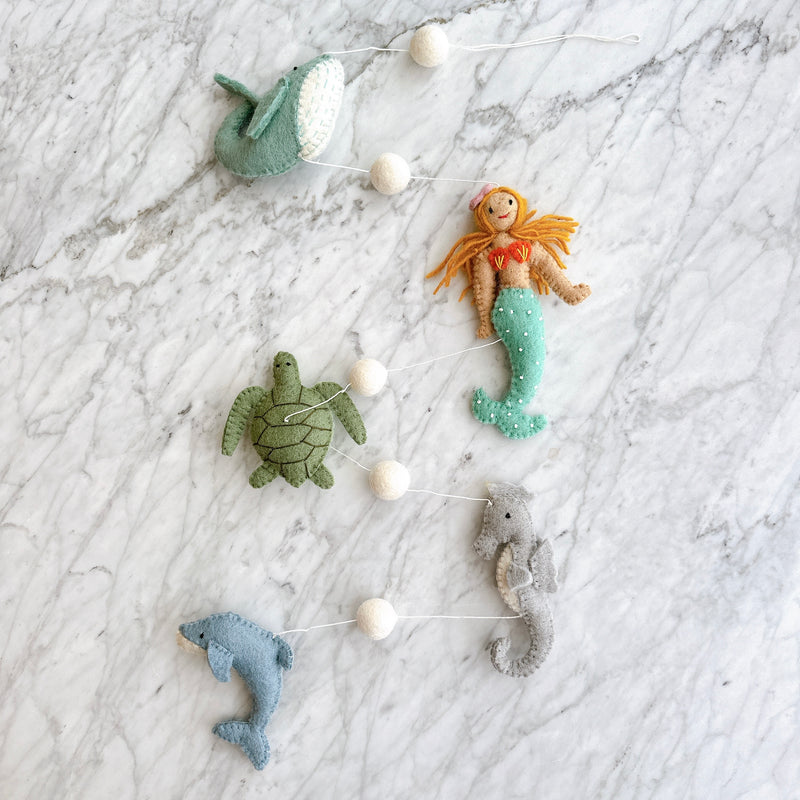 A Mermaid wool felt garland rests on a white marble surface, creating a whimsical scene. This felt garland features detailed wool felt figures including a light blue whale, a green sea turtle, a light blue dolphin, a grey seahorse, and a mermaid with orange hair and a green tail with white dots. White felt balls are interspersed between each figure along a horizontal white string.
