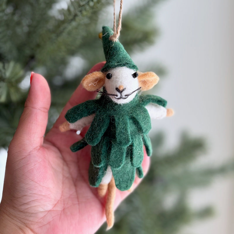 A wool felt Merry Spruce Mouse Christmas ornament is gently held by a female hand against a softly blurred Christmas tree background. The charming mouse ornament is dressed in a layered green costume, designed to mimic a Christmas tree, and wears a pointy elf-like hat. It features a white face with black eyes and whiskers, and is suspended by a jute string, highlighting its festive, handcrafted details.