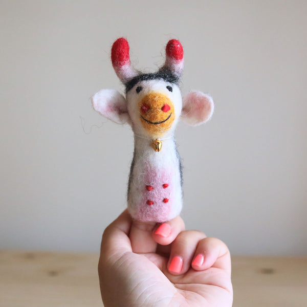 A needle felted milk cow finger puppet is held by a female hand against a neutral background. This handcrafted wool felt finger puppet features a white and gray body with red felt spots and a golden bell around its neck. The cow's head has red-tipped horns, a tan smiling muzzle, and floppy pink ears, creating a whimsical and playful design for imaginative play.