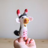 A handmade wool felt milk cow finger puppet rests on a fair-skinned fingertip with pink nails, set against a light background and wood surface. This intricately needle felted finger puppet features a white and grey body with red spots, a tiny golden bell around its neck, and red-tipped horns. Detailed craftsmanship is evident in its black and white striped head patch and small black eyes, making it a unique felt toy for kids.