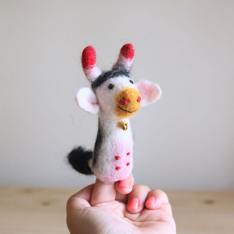A handmade wool felt milk cow finger puppet rests on a fair-skinned fingertip with pink nails, set against a light background and wood surface. This intricately needle felted finger puppet features a white and grey body with red spots, a tiny golden bell around its neck, and red-tipped horns. Detailed craftsmanship is evident in its black and white striped head patch and small black eyes, making it a unique felt toy for kids.
