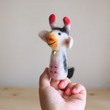 A handmade wool felt milk cow finger puppet is held by a female hand with coral-painted nails, resting on a light-colored wooden surface. This charming, fair-trade puppet, crafted from 100% natural wool felt in Nepal, features a white body with black markings and a black tail. It has distinct red-tipped horns, a light orange muzzle with red spots, and a gold bell around its neck, showcasing unique design and inviting imaginative play.