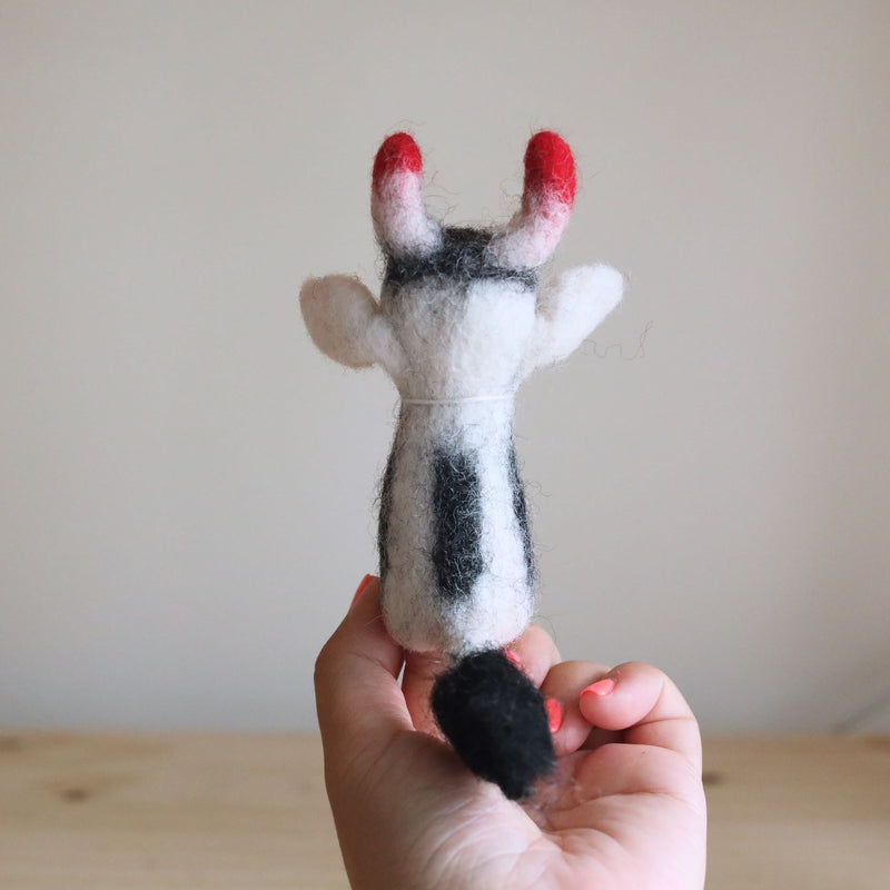 A wool felt milk cow finger puppet is held by a female hand against a light background, showcasing its details. This handcrafted felt kids toy features a white body with distinct black spots, a black tail, and small red-tipped horns.