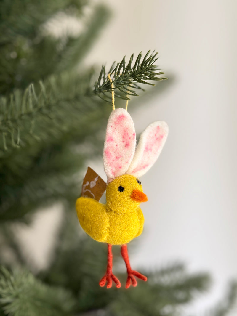 A whimsical handcrafted felt mini bunny chick ornament hangs from a Christmas tree branch. Its bright yellow chick body has an orange beak and detailed red felt legs. Long, soft white bunny ears with pink speckles top its head, complemented by a tan felt tail. It hangs from a yellow cotton string.