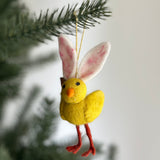 A playful Mini Bunny Chick ornament hangs from a pine tree branch against a bright background. This charming ornament is predominantly bright yellow, detailed with long, soft pink bunny ears, a small orange beak, and red legs. A gold string is attached for easy hanging.