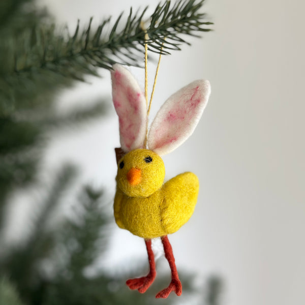 A playful Mini Bunny Chick ornament hangs from a pine tree branch against a bright background. This charming ornament is predominantly bright yellow, detailed with long, soft pink bunny ears, a small orange beak, and red legs. A gold string is attached for easy hanging.