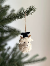 A cheerful wool felt graduate ram ornament hangs from a pine branch on a Christmas tree. This handmade felt ornament features curled brown horns, a fluffy cream coat, and a tiny black graduation cap, suspended by a jute string in a bright, festive setting.