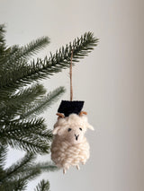 A handmade wool felt Graduate Ram ornament hangs from a green Christmas tree branch against a light background. This unique Christmas ornament features a cream-colored, fluffy body, small brown curled horns, and a tiny black graduation cap, complete with a natural twine string for hanging.