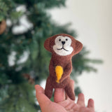 A handcrafted wool felt monkey finger puppet is held gently in a female hand, positioned in front of a softly blurred Christmas tree. This brown, needle-felted monkey features a white face with stitched black eyes and a smile, holding a vibrant yellow felt banana. Its charming, handmade design evokes playful creativity.