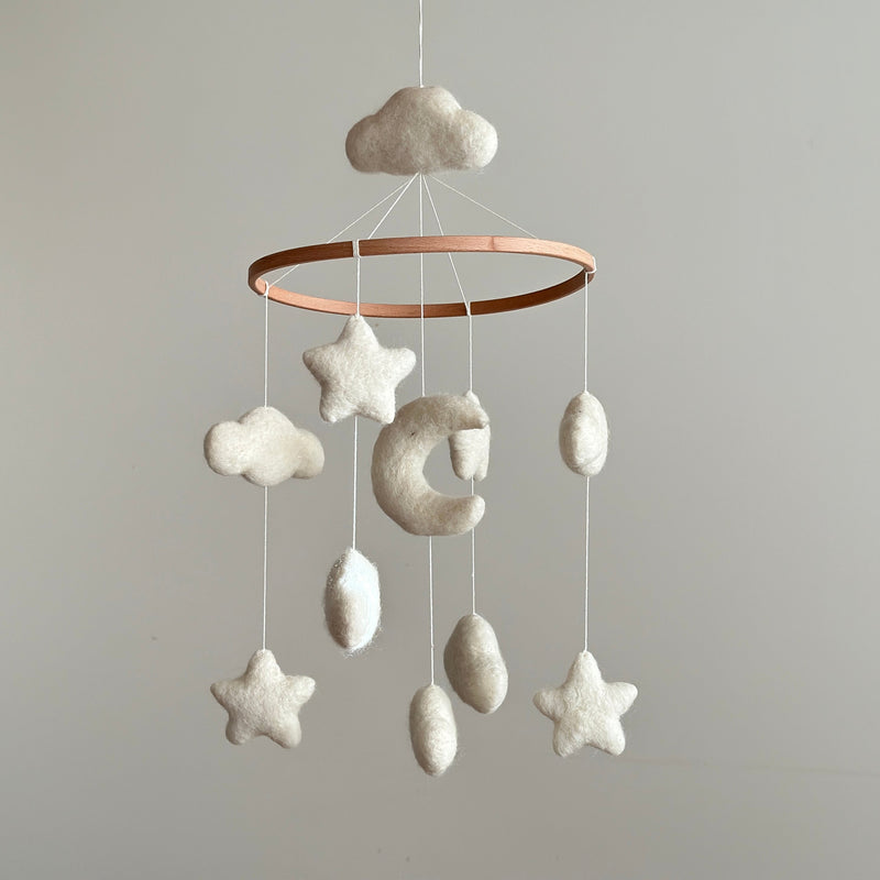 A dreamy Moon & Stars Baby Mobile hangs suspended against a plain light gray backdrop, crafted to evoke a sense of calm and wonder. It features soft, neutral-toned wool felt cloud, star, and crescent moon ornaments, delicately suspended from a natural wood hoop by strings. The wool felt clouds have a fluffy texture, while the stars are neatly shaped with pointed edges and the moon is crescent-shaped, creating a visually comforting and aesthetically pleasing product.